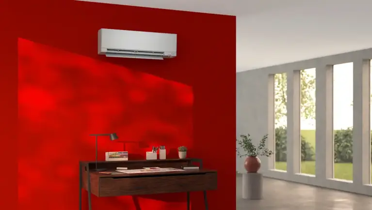 Weiße Wandklimaanlage an einer kräftig roten Wand über einem dunklen Holzschreibtisch. Rechts daneben sorgen bodentiefe Fenster für viel Tageslicht und Blick ins Grüne.