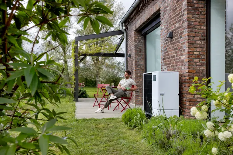 Außeneinheit einer Wärmepumpe an einer Backsteinfassade neben einer Holzterrasse. Eine Person sitzt an einem roten Gartentisch im begrünten Garten eines Einfamilienhauses.
