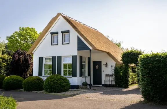 Frontalansicht des reetgedeckten Ferienhauses „Beach Cottage“ mit weißer Fassade, schwarzen Fensterläden und gepflegtem Garten