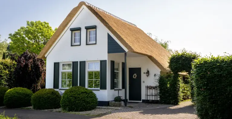 Frontalansicht des reetgedeckten Ferienhauses „Beach Cottage“ mit weißer Fassade, schwarzen Fensterläden und gepflegtem Garten