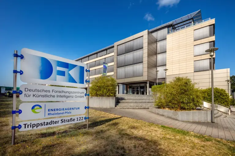 Modernes Büro- und Forschungsgebäude mit mehrstöckiger Fassade aus Glas und hellen Paneelen unter blauem Himmel; im Vordergrund ein großer weißer Firmenschild-Aufsteller mit den Logos von DFKI, der EnergieAgentur Rheinland-Pfalz sowie der Adresse „Trippstadter Straße 122“.