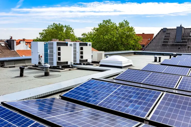 Plat dak van een commercieel gebouw met meerdere zonnepanelen en buitenunits van warmtepompen of airconditioningsystemen. Op de achtergrond zijn omliggende gebouwen en groene bomen zichtbaar onder een heldere blauwe lucht.