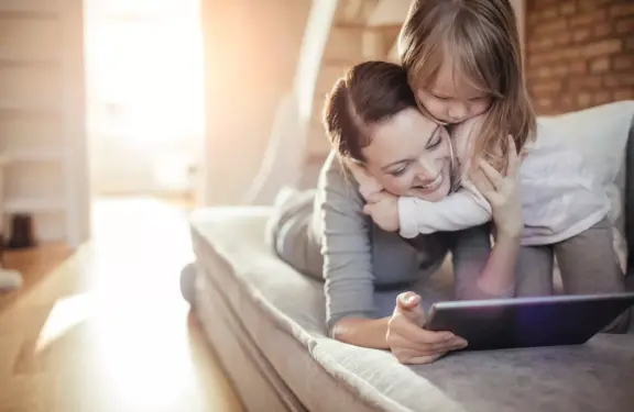 Une fille serre sa mère dans ses bras alors qu'elles sont assises ensemble sur le canapé et regardent une tablette.