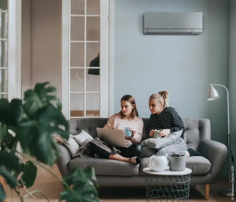 2 jonge meisjes zitten in de zetel met elk een beker in de hand, kijkend naar een laptop-scherm. Op de muur achter hun hangt een grijze MSZ-EF Wandunit