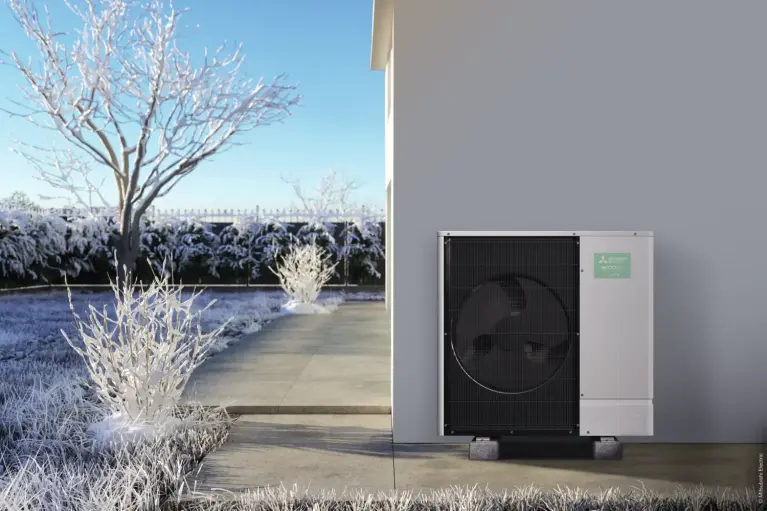 Buitenunit van een lucht-water warmtepomp geplaatst naast een moderne woning. De tuin en planten zijn bedekt met sneeuw, terwijl de warmtepomp zorgt voor energiezuinige verwarming en warm water tijdens de winter.