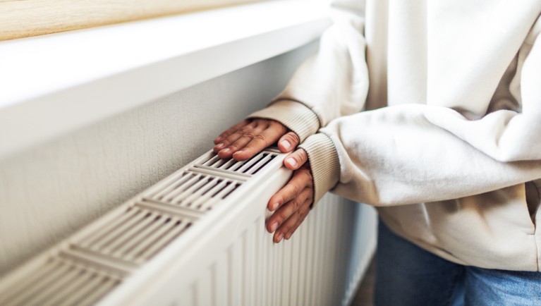 Persoon houdt de handen boven een radiator onder een venster om de warmte te voelen. De radiator is geplaatst tegen een lichte muur in een woning.
