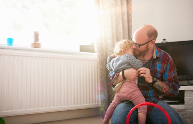 Persoon zit op de vloer naast een radiator en houdt een klein kind vast in een warme en lichte woonkamer. Daglicht valt binnen via het raam boven de radiator.