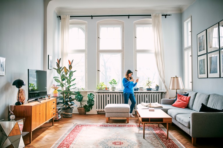 Ruime en lichte woonkamer met hoge ramen, planten en een radiator onder de vensters. Een persoon staat in de leefruimte met een tablet, terwijl natuurlijk daglicht de kamer verlicht.