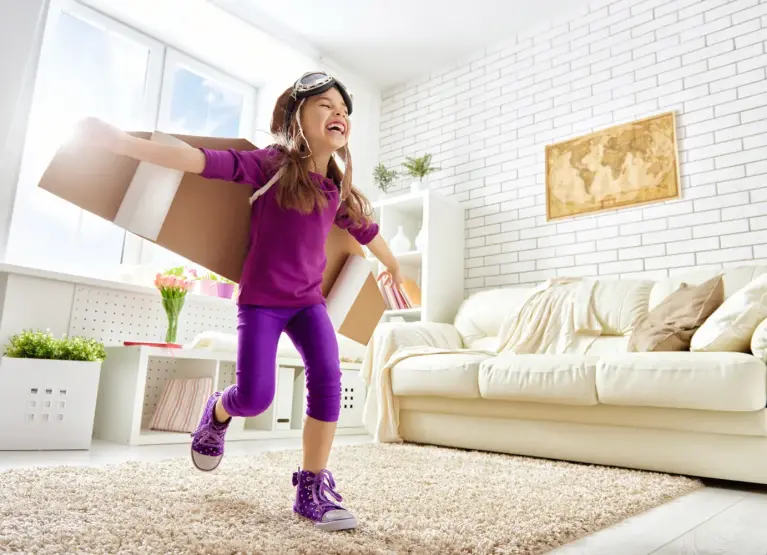 Een klein meisje met paarse kleding aan staat in een woonkamer met een kartonnen vlieger op haar rug.