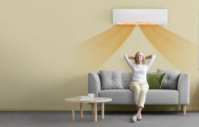 Wandgemonteerde airconditioner verspreidt warme lucht in een moderne woonkamer. Een persoon zit op een grijze zetel onder de unit.