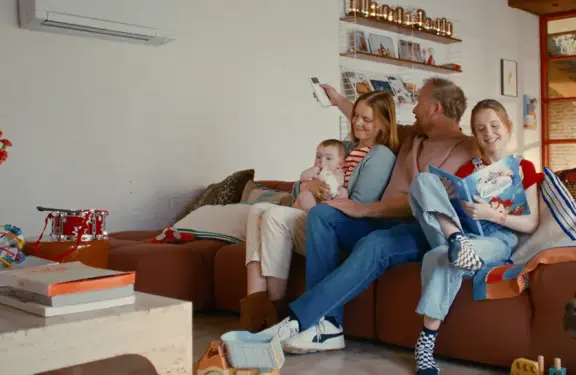 Gezin zit in de zetel in de living en de papa zet de airco aan met de afstandbediening